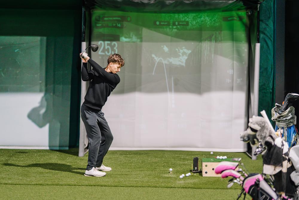 Golfer driving in an indoor golf simulator bay with launch data on the screen