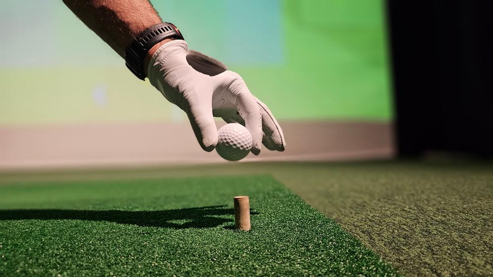 Gloved hand placing a golf ball on a tee in a simulator hitting mat